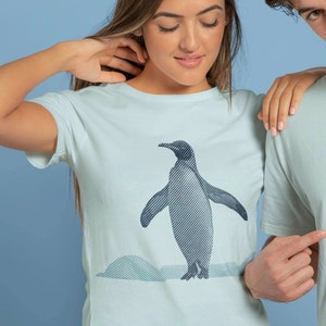 Könnte beinhalten: Ein hellblaues T-Shirt mit einem Aufdruck eines Pinguins, der auf einer schneebedeckten Landschaft steht. Der Pinguin ist in Schwarz-Weiß mit Streifen umrandet.