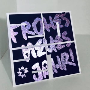 Puede incluir: Una tarjeta negra con un dise&ntilde;o de l&aacute;mina plateada que dice "Frohes Neues Jahr!" en una fuente estilizada.