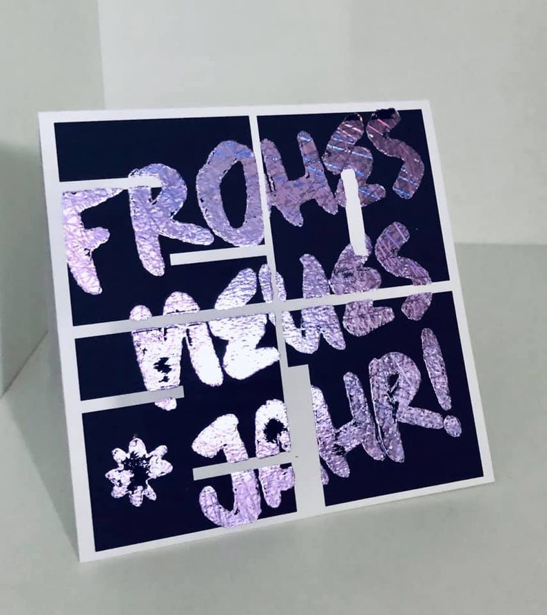 Puede incluir: Una tarjeta negra con un dise&ntilde;o de l&aacute;mina plateada que dice "Frohes Neues Jahr!" en una fuente estilizada.