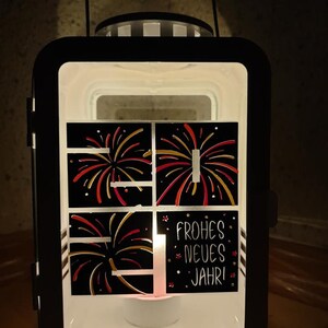 Puede incluir: Linterna de metal negro con una ventana de vidrio que presenta un dise&ntilde;o de fuegos artificiales rojos y dorados. La linterna tiene una vela de t&eacute; blanca en el interior y el texto "Frohes Neues Jahr!" en el vidrio.
