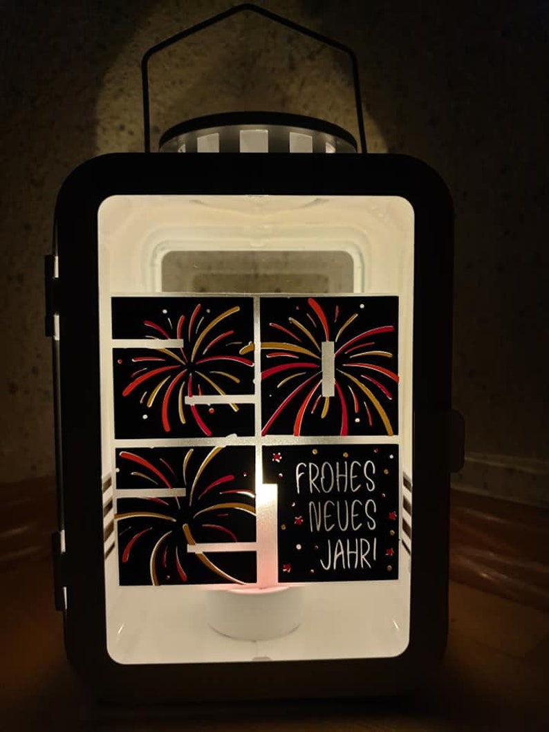 Puede incluir: Linterna de metal negro con una ventana de vidrio que presenta un dise&ntilde;o de fuegos artificiales rojos y dorados. La linterna tiene una vela de t&eacute; blanca en el interior y el texto "Frohes Neues Jahr!" en el vidrio.
