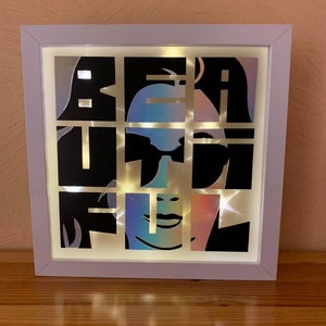 Könnte beinhalten: Eine weiße, gerahmte Lichtbox mit dem Wort "BEAUTIFUL" in schwarzen Buchstaben. Die Buchstaben sind ausgeschnitten, um ein Gesicht einer Frau mit Sonnenbrille hinter den Buchstaben zu zeigen. Die Lichtbox wird mit warmweißen LED-Lichtern beleuchtet.