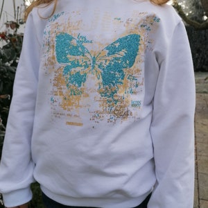 Op de afbeelding: Witte longsleeve trui met een turquoise vlinderprint en gouden glitteraccenten. De tekst "AEIOS 1990s" is op de trui gedrukt.