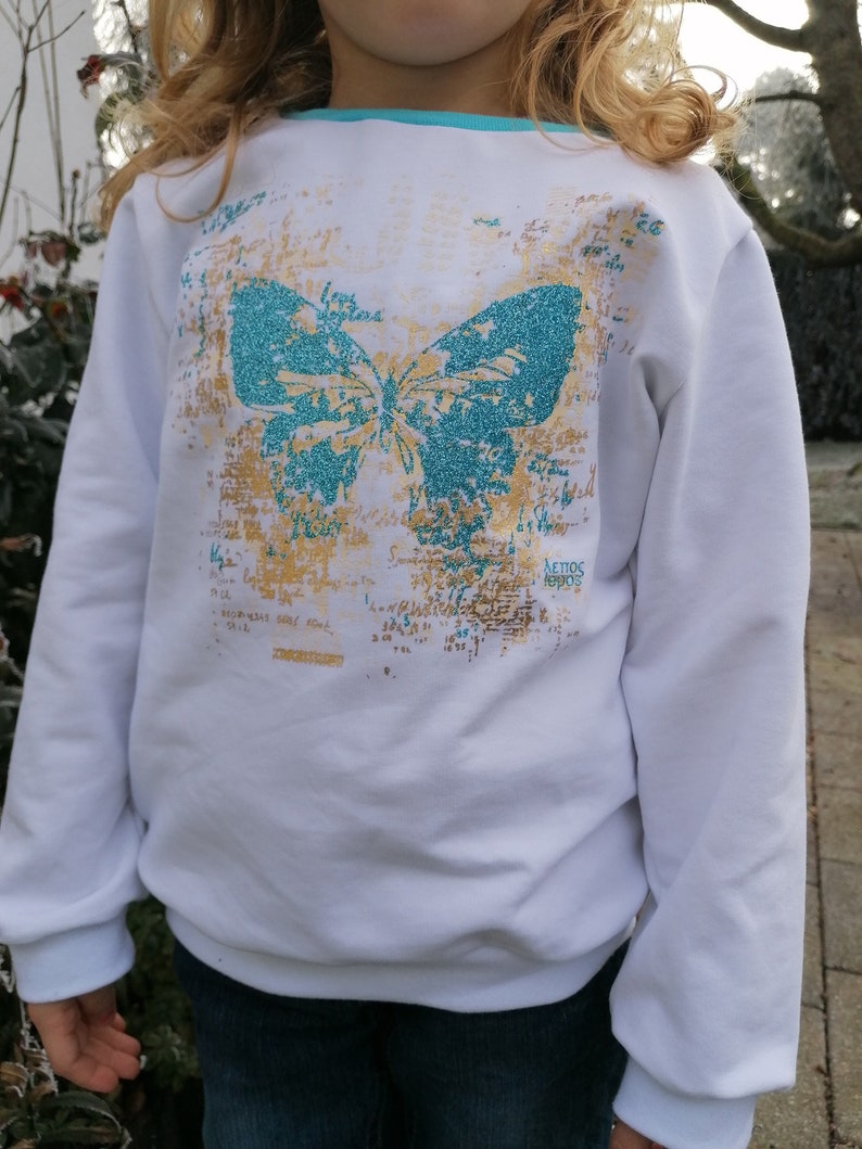 Op de afbeelding: Witte longsleeve trui met een turquoise vlinderprint en gouden glitteraccenten. De tekst "AEIOS 1990s" is op de trui gedrukt.