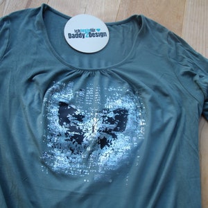 Op de afbeelding: Een tealgroen t-shirt met een zilveren vlinderprint. De vlinder is gemaakt van pailletten en heeft zwarte accenten. Het shirt heeft een ronde hals en korte mouwen. De tekst "Ich teste f&uuml;r Daddy2Design" staat op een witte ronde sticker.