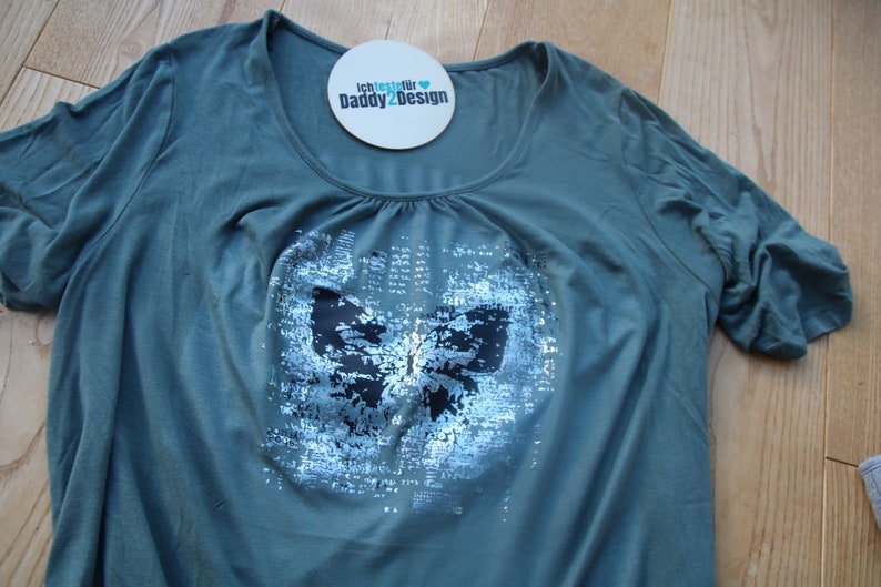 Op de afbeelding: Een tealgroen t-shirt met een zilveren vlinderprint. De vlinder is gemaakt van pailletten en heeft zwarte accenten. Het shirt heeft een ronde hals en korte mouwen. De tekst "Ich teste f&uuml;r Daddy2Design" staat op een witte ronde sticker.