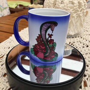 Könnte beinhalten: Eine blaue und weiße Keramiktasse mit einem Design aus grüner Schlange und roten Rosen. Die Tasse hat einen blauen Henkel und steht auf einem schwarzen drehbaren Präsentationsständer.