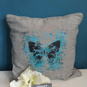 Op de afbeelding: Een grijs kussen met een blauw en zwart vlinderontwerp. De vlinder is omgeven door een verweerde, textuurrijke achtergrond met het woord "Lepidoptera" in zwart gedrukt. Het kussen ligt op een blauwe achtergrond.