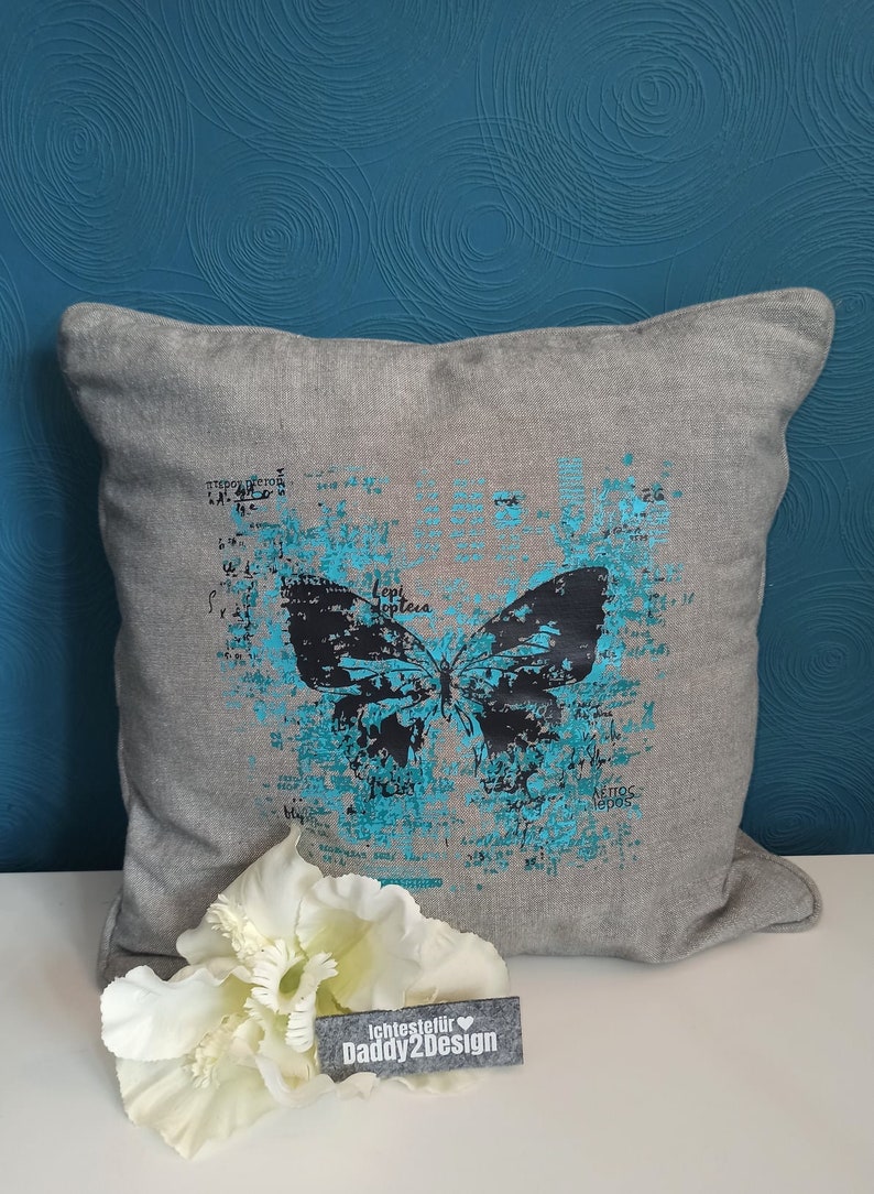 Op de afbeelding: Een grijs kussen met een blauw en zwart vlinderontwerp. De vlinder is omgeven door een verweerde, textuurrijke achtergrond met het woord "Lepidoptera" in zwart gedrukt. Het kussen ligt op een blauwe achtergrond.