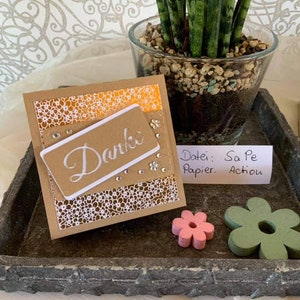 Op de afbeelding: Een bruine kaart met een wit kantpatroon en het woord "Danke" in wit op een wit rechthoek. De kaart ligt op een grijze stenen dienblad met een kleine roze bloem en een groene bloem.