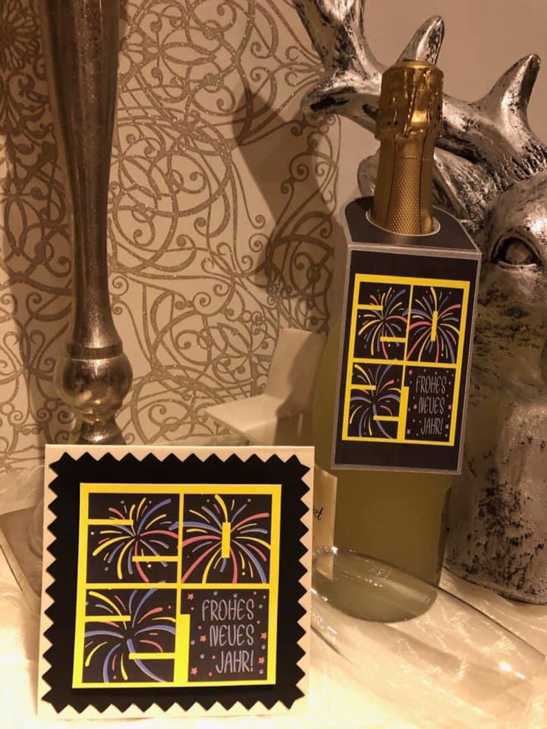 Puede incluir: Una tarjeta negra y amarilla con un dise&ntilde;o festivo de fuegos artificiales y el texto "Frohes Neues Jahr!" La tarjeta est&aacute; sobre una mesa junto a una botella de champ&aacute;n.