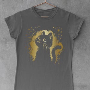 Op de afbeelding: Een grijs t-shirt met een gouden illustratie van een kat met een pluizige manen en staart. Het gezicht van de kat is gedetailleerd met snorharen en ogen. Gouden sterren en stippen omringen het kattenontwerp. Het shirt hangt aan een koperkleurige hanger.