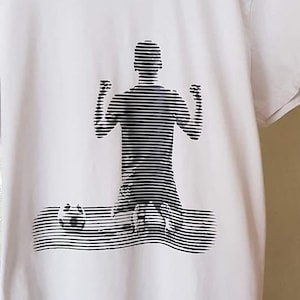 May include: A white t-shirt featuring a black and white graphic of a person kneeling with their arms raised.