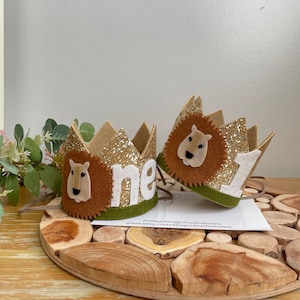 May include: Two gold glitter lion crowns with brown felt lion faces and the word "one" in white felt. The crowns have green felt trim.