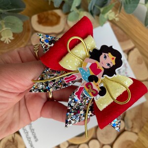 Puede incluir: Un lazo de brillo rojo y amarillo con un personaje de Wonder Woman en el centro. El lazo tiene un alambre dorado envuelto alrededor.