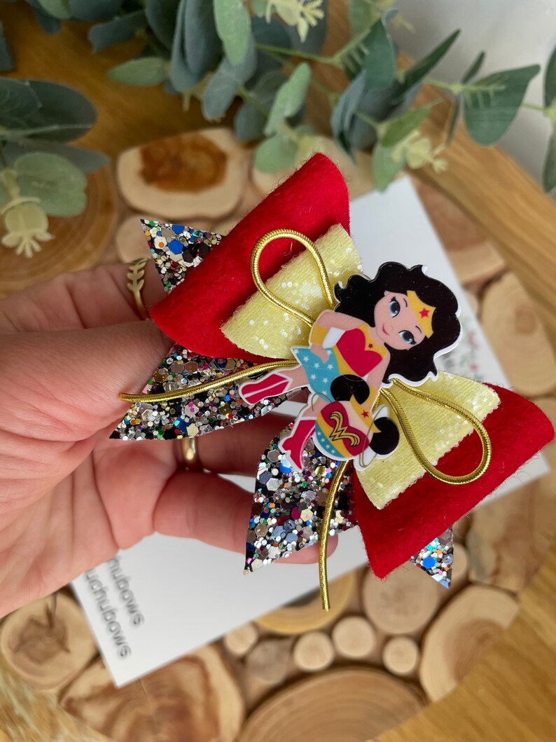 Puede incluir: Un lazo de brillo rojo y amarillo con un personaje de Wonder Woman en el centro. El lazo tiene un alambre dorado envuelto alrededor.