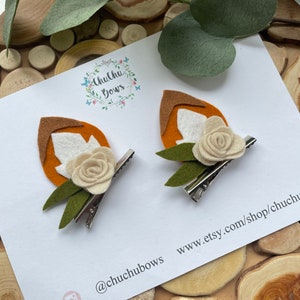 May include: Two felt fox hair clips with orange and white felt, green felt leaves, and a white felt flower. The clips are on a white card with the text "Chuchu Bows" and "www.etsy.com/shop/chuchubows" and the social media handles "@chuchubows", "f", and "instagram".