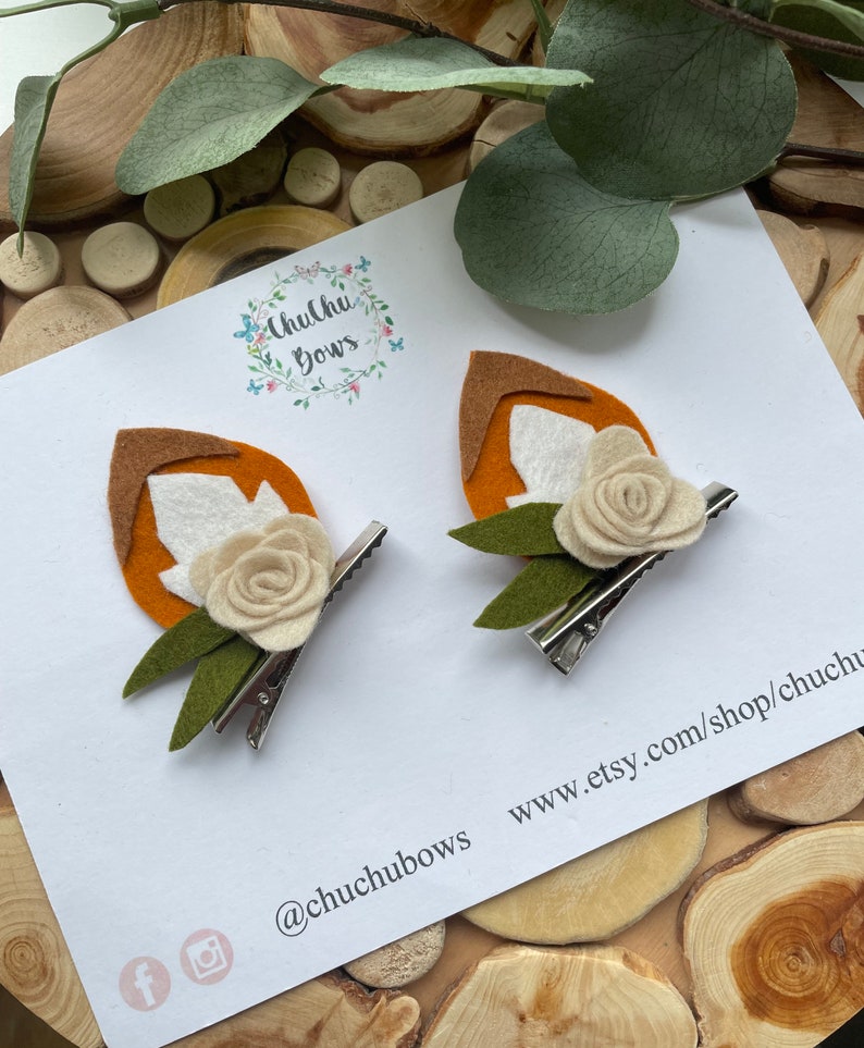 May include: Two felt fox hair clips with orange and white felt, green felt leaves, and a white felt flower. The clips are on a white card with the text "Chuchu Bows" and "www.etsy.com/shop/chuchubows" and the social media handles "@chuchubows", "f", and "instagram".