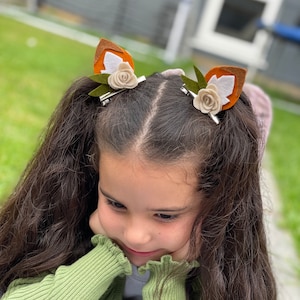 May include: A young girl with long brown hair wearing a green sweater and two brown and white felt fox ear hair clips with white flowers.