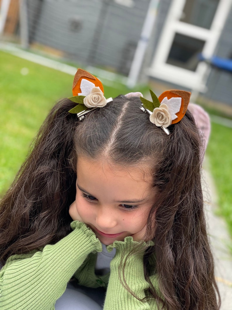 May include: A young girl with long brown hair wearing a green sweater and two brown and white felt fox ear hair clips with white flowers.