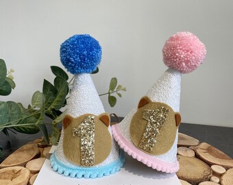 May include: Two white party hats with glitter, brown felt bear ears, and gold glitter number 1. One hat has a blue pom-pom and blue trim, the other has a pink pom-pom and pink trim.  Instagram: @chuchubows www.etsy.com/shop/chuchubows
