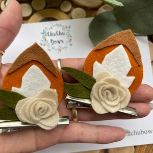 May include: Two felt hair clips with a brown, orange and white leaf design. Each clip has a white felt flower with a green felt leaf.