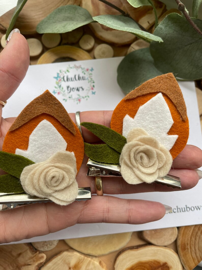 May include: Two felt hair clips with a brown, orange and white leaf design. Each clip has a white felt flower with a green felt leaf.