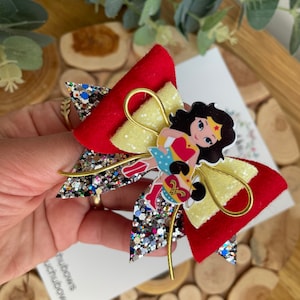 Puede incluir: Un lazo de brillo rojo, amarillo y plateado con un personaje de Wonder Woman en el centro. El lazo est&aacute; atado con una cinta dorada.