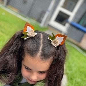 May include: A young girl with long brown hair wearing two brown and white felt fox ear hair clips with green leaves and white flowers.