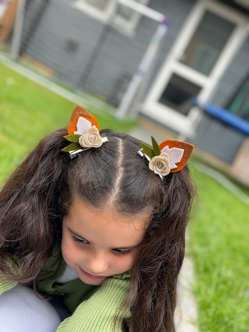 May include: A young girl with long brown hair wearing two brown and white felt fox ear hair clips with green leaves and white flowers.