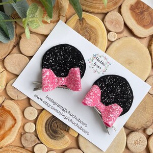 Puede incluir: Dos orejas de Minnie Mouse de purpurina negra con lazos de purpurina rosa unidos a pinzas de cocodrilo plateadas. Las orejas est&aacute;n sobre un fondo blanco con el texto "Chuchu Bows" y "Instagram@chuchubows www.etsy.com/shop/chuchubows".