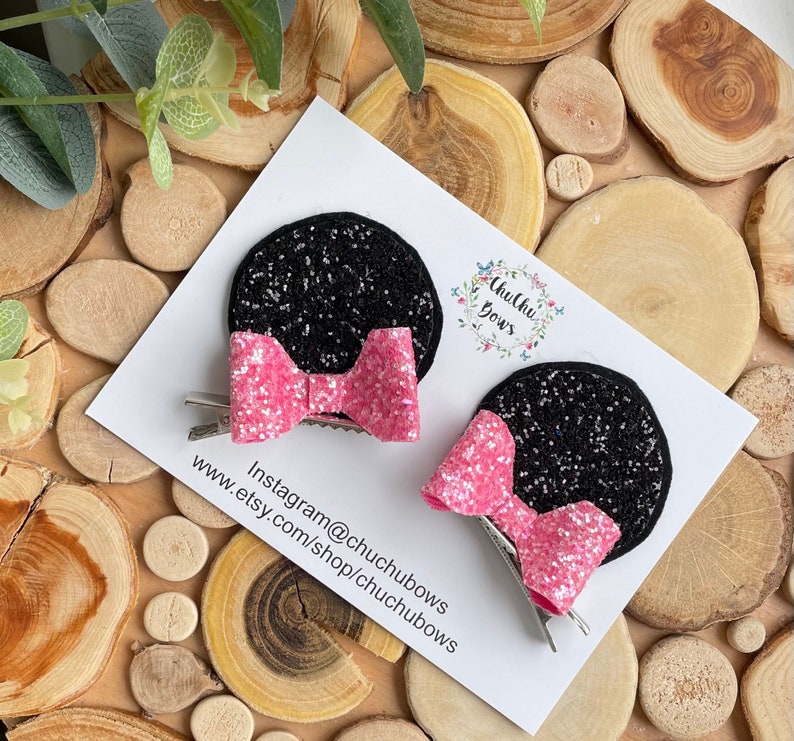 Puede incluir: Dos orejas de Minnie Mouse de purpurina negra con lazos de purpurina rosa unidos a pinzas de cocodrilo plateadas. Las orejas est&aacute;n sobre un fondo blanco con el texto "Chuchu Bows" y "Instagram@chuchubows www.etsy.com/shop/chuchubows".