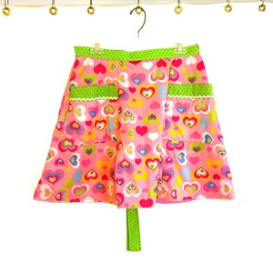 May include: A pink apron with a green trim and a heart pattern. The apron has two pockets and a green tie.
