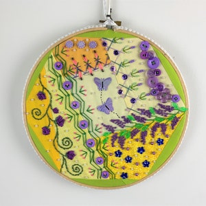 May include: Embroidered wall art in a wooden hoop. The design features yellow, green, and lavender fabric sections with floral and butterfly motifs. Embellishments include purple buttons, beads, and embroidery stitches. A decorative piece for home decor.