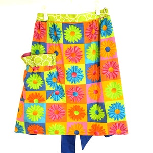 May include: A colorful apron with a large pocket and a bright blue bow. The apron is made of a fabric with a checkered pattern of large, colorful daisies.