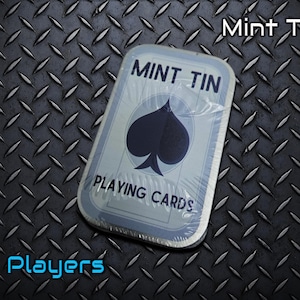May include: A rectangular mint tin containing playing cards, wrapped in clear plastic. The tin features the words "MINT TIN" and "PLAYING CARDS" with a black spade symbol. The background is a textured, dark gray metal.