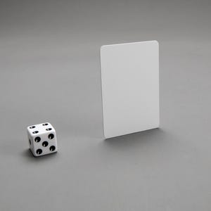 Puede incluir: Un dado blanco con puntos negros en cada lado, sentado en una superficie gris junto a una tarjeta blanca.