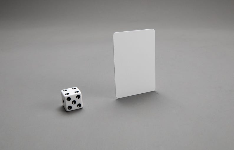 Puede incluir: Un dado blanco con puntos negros en cada lado, sentado en una superficie gris junto a una tarjeta blanca.