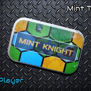 Può includere: Una scatola di menta rettangolare con la scritta "Mint Knight" in un font pixelato. La scatola ha un design colorato con pixel art verde, blu e giallo. Il testo "Mint Tin!" è sopra e "1 Player" sotto.