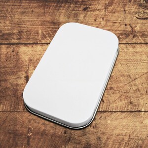May include: A white rectangular tin with rounded corners, set against a wooden background. The tin has a silver rim and a smooth, blank surface, suggesting it's designed for customization or storage. The wood grain adds a rustic contrast.