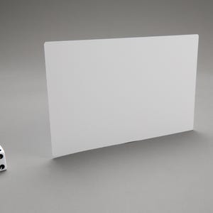 Puede incluir: Un dado blanco de seis caras con puntos negros en cada cara, sentado sobre una superficie gris frente a una tarjeta rectangular blanca.