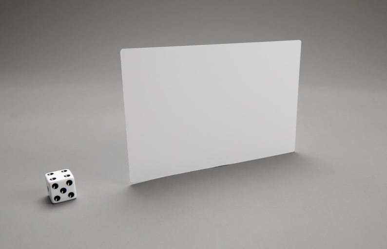 Puede incluir: Un dado blanco de seis caras con puntos negros en cada cara, sentado sobre una superficie gris frente a una tarjeta rectangular blanca.