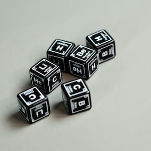 May include: Close-up of six black dice with white lettering. Each die features a different element symbol and name, including Helium, Lithium, Beryllium, Carbon, and Boron.