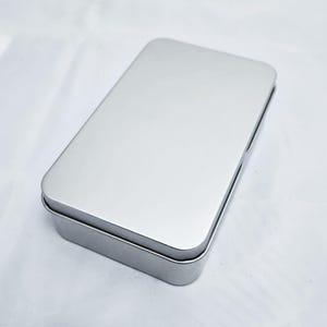May include: A rectangular, silver-colored metal tin with a hinged lid. The tin is empty and sits on a white surface. The lid is closed, and the edges of the tin are slightly rounded.
