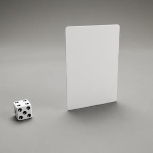 Puede incluir: Un dado blanco con puntos negros sobre un fondo gris. Una tarjeta blanca est&aacute; de pie detr&aacute;s del dado.