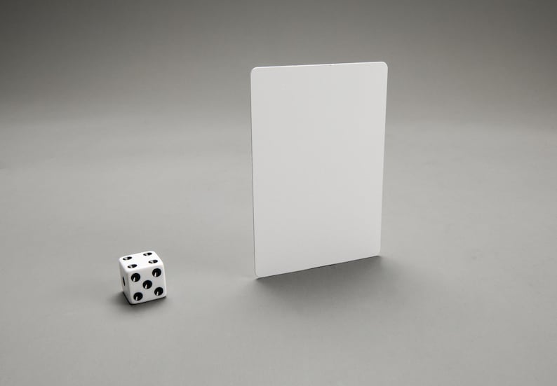 Puede incluir: Un dado blanco con puntos negros sobre un fondo gris. Una tarjeta blanca est&aacute; de pie detr&aacute;s del dado.