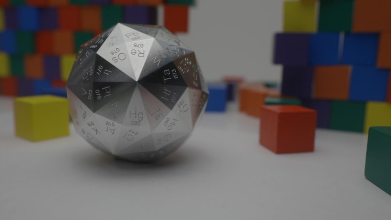 Pu&ograve; includere: Un dado dodecaedrico geometrico argentato con la tavola periodica degli elementi. Il dado &egrave; appoggiato su una superficie bianca, con blocchi di legno colorati sullo sfondo.