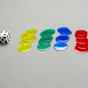 Puede incluir: Un dado blanco con puntos negros y cinco pilas de fichas de juego de colores. Las fichas son amarillas, verdes, azules y rojas.