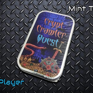 May include: A rectangular mint tin with the text "Crypt Crawler Quest" and a graphic of a snake, treasure, and weapons. The text "Mint Tin!" and "1 Player" are also visible. The tin is wrapped in clear plastic.