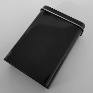 May include: A black rectangular tin with a glossy, reflective surface. The lid features a thin, silver-coloured metal trim. The tin is angled, highlighting its shiny finish. The design is simple and modern.